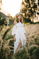 Fototapeta na wymiar A beautiful tender young girl in a white dress with a wreath of flowers on her head is standing in a spring field at sunset. Happy bride with blonde curly hair enjoying sunny day. Summer tender photo.