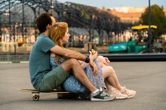 Back view of young couple sitting together on longboard against blurred cityscape and looking away while having lunch at daytime 