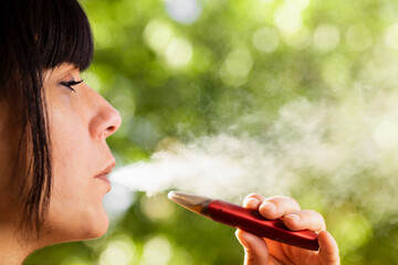 Close up of a woman exhaling vapor from an electronic cigarette in the street with nature in the...