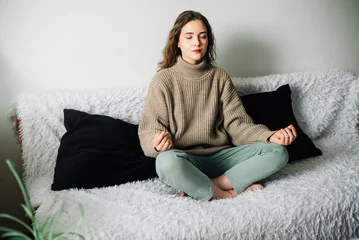 Fototapeten Young woman sitting in lotus position on the sofa with her hands on her knees, meditating, trying to relax her mind and relieve stress © Yauhen