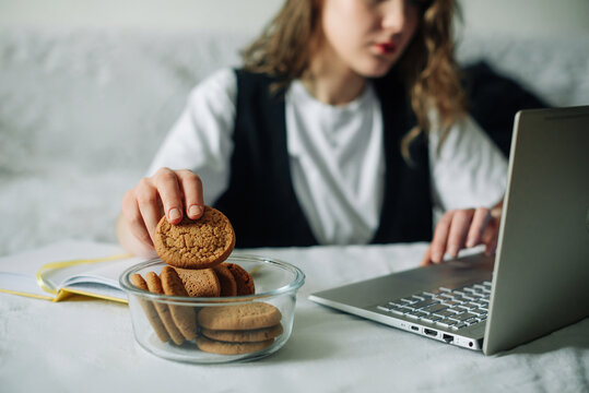 Unconscious eating. Bad habits. Selective focus of hand of concentrated female working on laptop at the kitchen table, taking unintentionally and automatically cookies from a transparent glass jar
