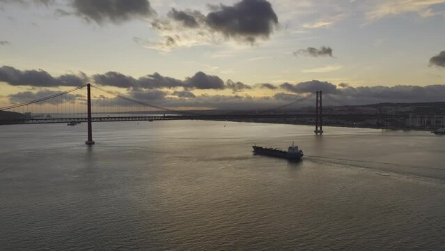 Drone shot rising and reversing away from a tanker ship heading under the bridge in lisbon.