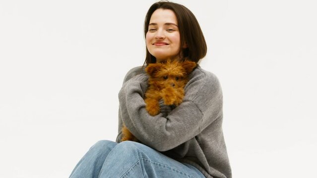 young beautiful woman in casual clothes hugs, kisses her beloved small fluffy dog while sitting on the floor on a white background. The concept of communicating with animals