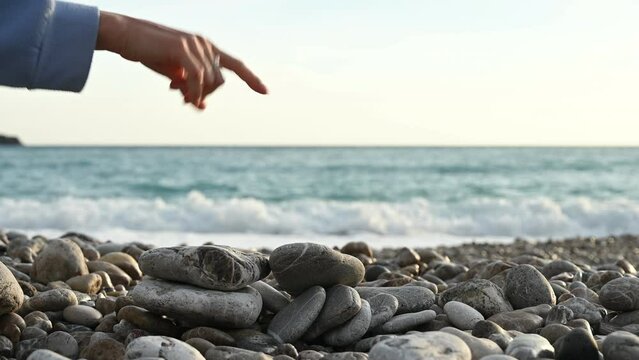 Woman ruins balanced rock pyramid on pebbles beach against sea waves on background