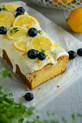 Homemade lemon pound cake decorated with cream cheese frosting, fresh lemon slices and blueberries on baking paper on cutting board.
