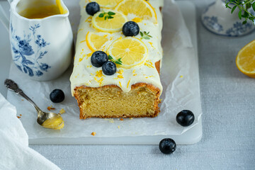 Homemade lemon pound cake decorated with cream cheese frosting, fresh lemon slices and blueberries on baking paper on cutting board and lemon curd in vintage saucer on linen tablecloth.