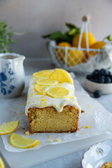 Homemade lemon pound cake decorated with cream cheese frosting, fresh lemon slices and blueberries on baking paper on cutting board and lemon curd in vintage saucer on linen tablecloth.