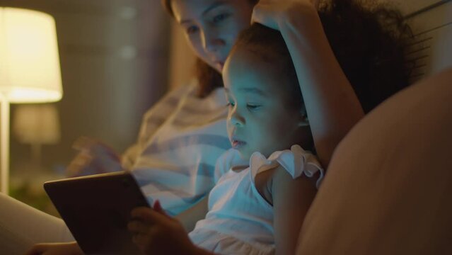Medium close up shot of Asian mother hugging lovely toddler daughter, stroking her head and using smartphone as girl watching cartoon on digital tablet on bed in evening at home