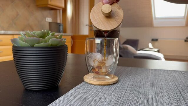 Pouring Cold Brew Coffee from French Press to Transparent Glass. Male Hand with Brown Liquid and Ice Cubes Inside at Home.