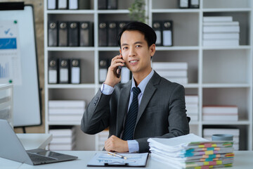 young Asian businessman involved in cellphone call conversation working on computer, giving professional consultation to client or negotiating project.