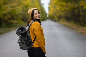 Cheerful young woman in yellow jumper smiles looking at camera. Pretty lady goes on hitchhiking trip around world. Traveller waits for passing car against yellow forest, copyspace