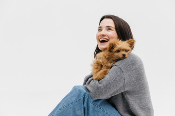 young beautiful woman in casual clothes hugs, kisses her beloved small fluffy dog while sitting on...