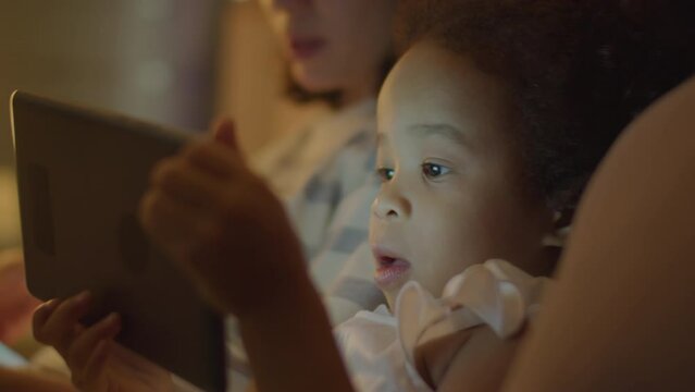Close up tilt down shot of Asian mother smiling at talking to cute toddler daughter as she watching cartoon on digital tablet in evening at home