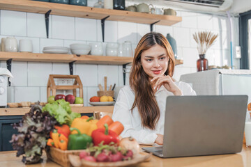 Young happy smiling woman using laptop while cooking vegan food in the modern kitchen, smiling hipster girl preparing healthy vitamin salad with vegetables, eating healthy lifestyle