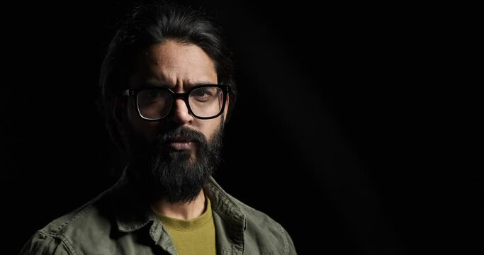Slow motion shot of bearded serious young man with eyeglasses looking angrily at camera isolated against black background