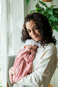 young mother with curly hair poses looking at camera while holding her newborn baby in her arms sucking his thumb. family and newborn care concept.