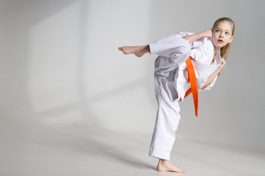 A strong little girl in a kimono kicks on a white background.