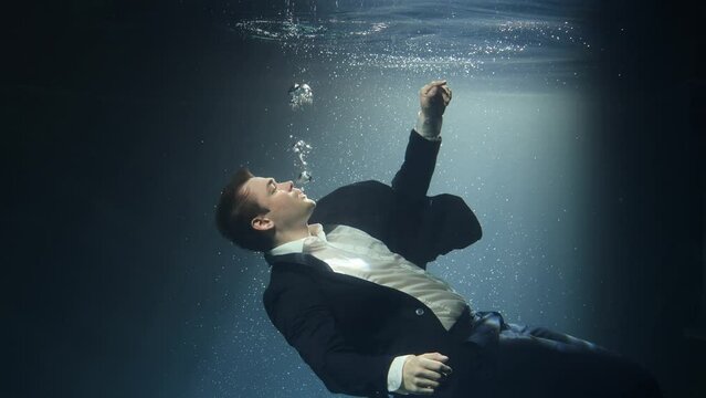 Businessman, a man in a suit, is drowning under water, reaching for the surface of the water.