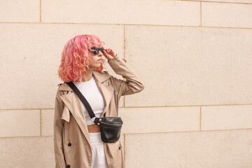 Portrait shooting of a stylish girl with pink hair. Beige shades. Trends of spring and summer 2023.Beige Trench cloak with white top and pants. Black small belt bag