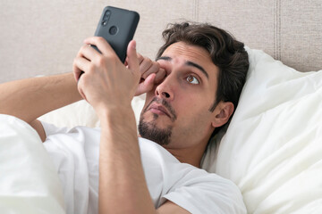 Surprised and shocked man lying on the bed and looking at smartphone at the morning time. In concept of bad news and checking up mobile after awakening