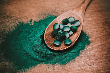 Green spirulina pills in light wooden spoon with spirulina algae powder at wooden background. Superfood and healthy detox diet.