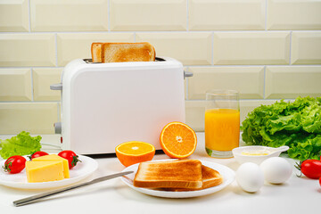 Healthy breakfast in the kitchen. Toaster and bread toast, eggs, tomatoes, freshly squeezed orange juice. Cheese, butter, lettuce. White toaster with healthy food and drinks on table in light kitchen