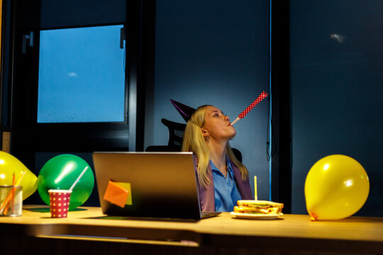 Upset woman in birthday party hat with playing pipe celebrating alone at workplace. Sad lovely businesswoman sitting at desktop blowing party whistle