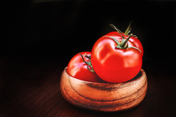 Red glossy ripe tomatoes in wooden bowl at black background. Dark luxury style vegetables.