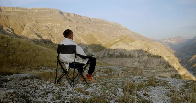 Young attractive hipster man sitting on a camping folding chair in a clearing against the backdrop of a mountainous dry southern landscape and looking into the distance. High quality 4k footage