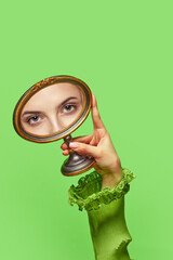 Female hand holding small mirror with reflection of adorable, green girl's eyes over bright green...