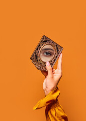 Female hand holding small mirror with reflection of beautiful, green girl's eye over orange...