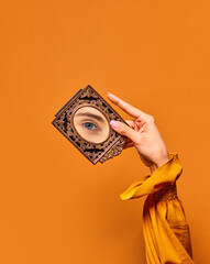 Female hand holding small mirror with reflection of girls eye over orange background. Eyesight....