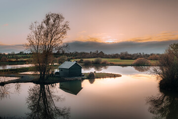 An idyllic image of a small fishing cabin on a tranquil island between two lakes, with the sunset and its reflections casting a warm glow on the water