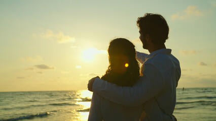 Standing side by side, a young couple gazes out at the sea with a deep sense of romance, their eyes fixed on the stunning sunset that paints the sky with a warm, orange glow. Slow motion shot.