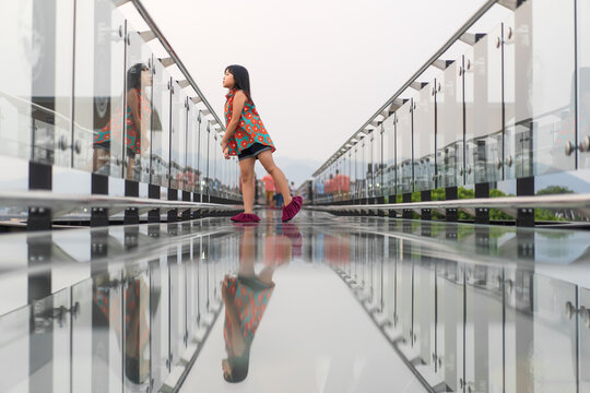 Asian child or kid girl happy standing on skywalk bridge with reflection and clear glass floor on mountain view and fear of heights for vacation travel and holiday relax at Kanchanaburi in Thailand