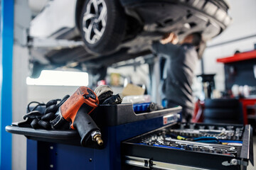 Selective focus on a drill and drawer with tools on a mechanic toolbox with worker fixing car in blurry background at mechanic's shop.