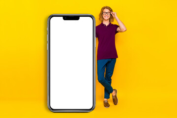 Full body photo of funny ginger guy stand near big telephone wear spectacles t-shirt jeans pants sneakers isolated on yellow background
