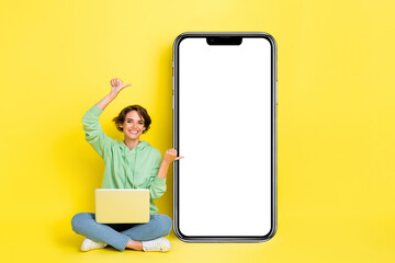 Full length photo of cheerful sweet girl dressed green sweatshirt pointing thumbs modern device empty space isolated yellow color background