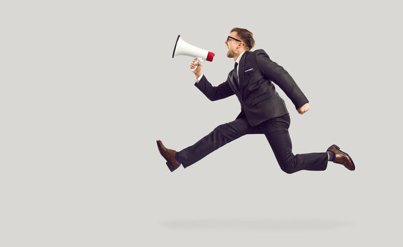 Crazy funny man in suit and glasses isolated on light gray background jumping high in air, yelling through megaphone, making announcement, advertising vacancy, or drawing attention to urgent problem