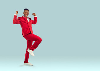 Fototapeta na wymiar Happy emotional stylish african american man in suit sincerely rejoices in his success isolated on pastel light blue background. Joyful man in red suit happily clenching his fists near copy space.