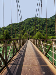 Obraz premium Suspension metal automobile bridge in the mountains over the river to the Georgian village. Green forest in the mountains in the background. No people. copy space