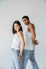 Fototapeta premium young and trendy interracial couple in white tank tops and jeans looking at camera isolated on grey.