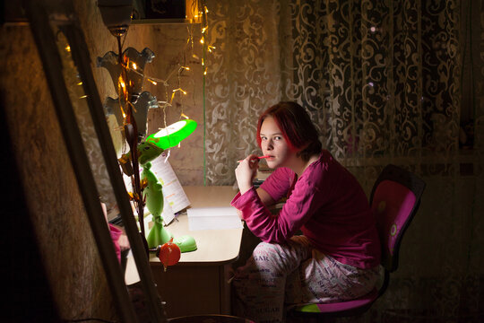 A schoolgirl girl with red hair sits at a table with a table lamp and a luminous garland, reads a book, does her homework, in a dark room in the evening. Image about studying at home.