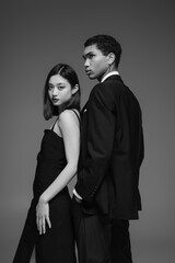 sensual asian woman in black strap dress looking at camera near african american man in elegant suit isolated on grey.