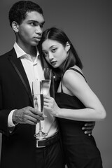 young and fashionable interracial couple in formal wear holding champagne glasses isolated on grey.