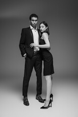 full length of african american man in elegant suit and asian woman in black dress looking at camera on grey background.