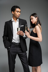 elegant african american man in suit and asian woman in black dress posing with champagne glasses isolated on grey.