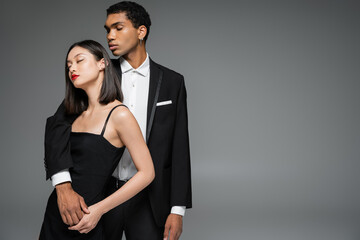stylish african american man in elegant suit embracing asian woman posing with closed eyes isolated on grey.