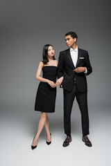 full length of interracial couple in black elegant attire looking at each other on grey background.