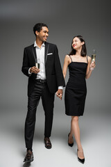 full length of elegant multiethnic couple with champagne glasses holding hands and smiling at each other while walking on grey background.